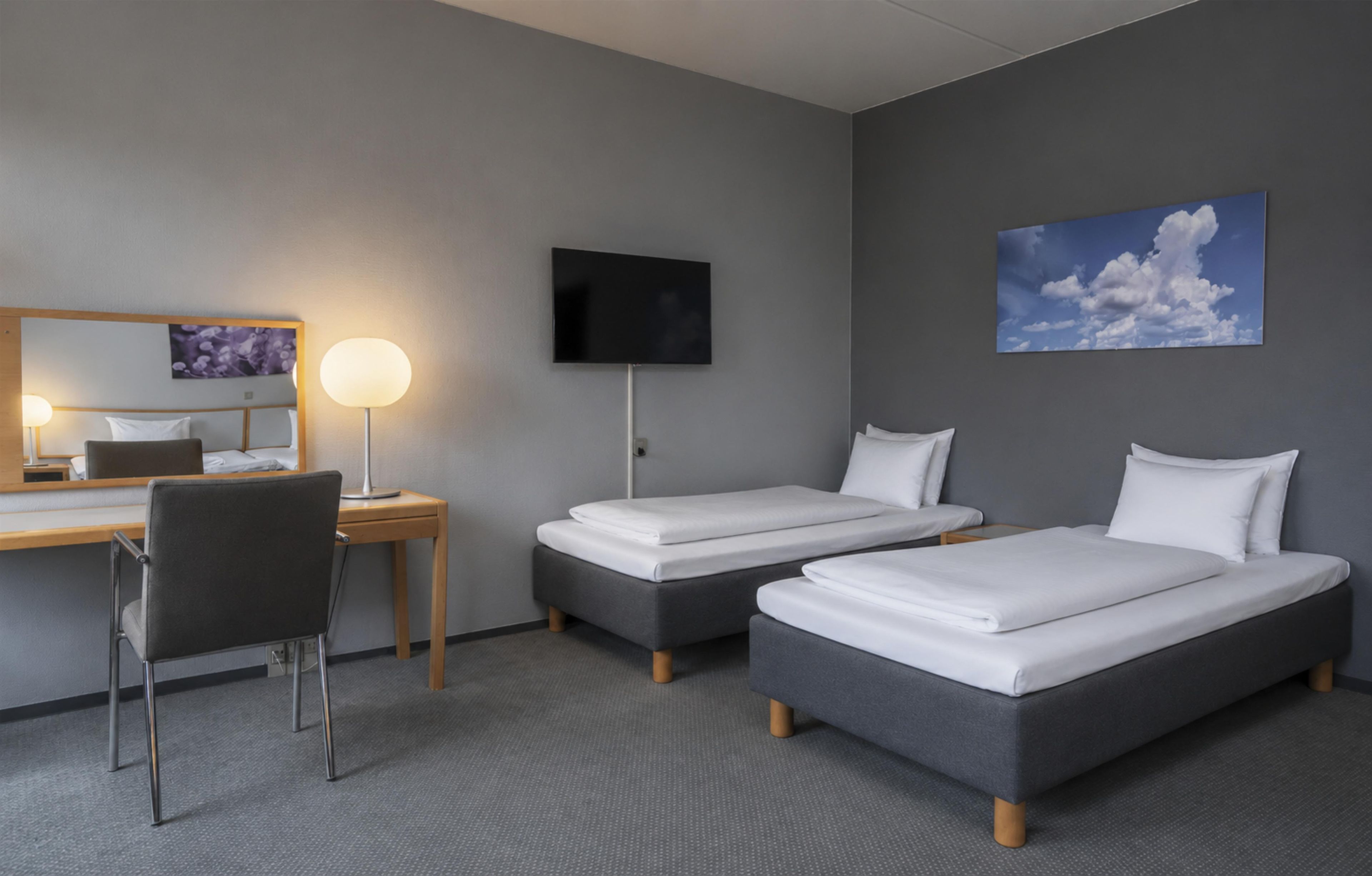 A hotel room with two beds, a desk, a chair, a TV, and a painting on the wall.