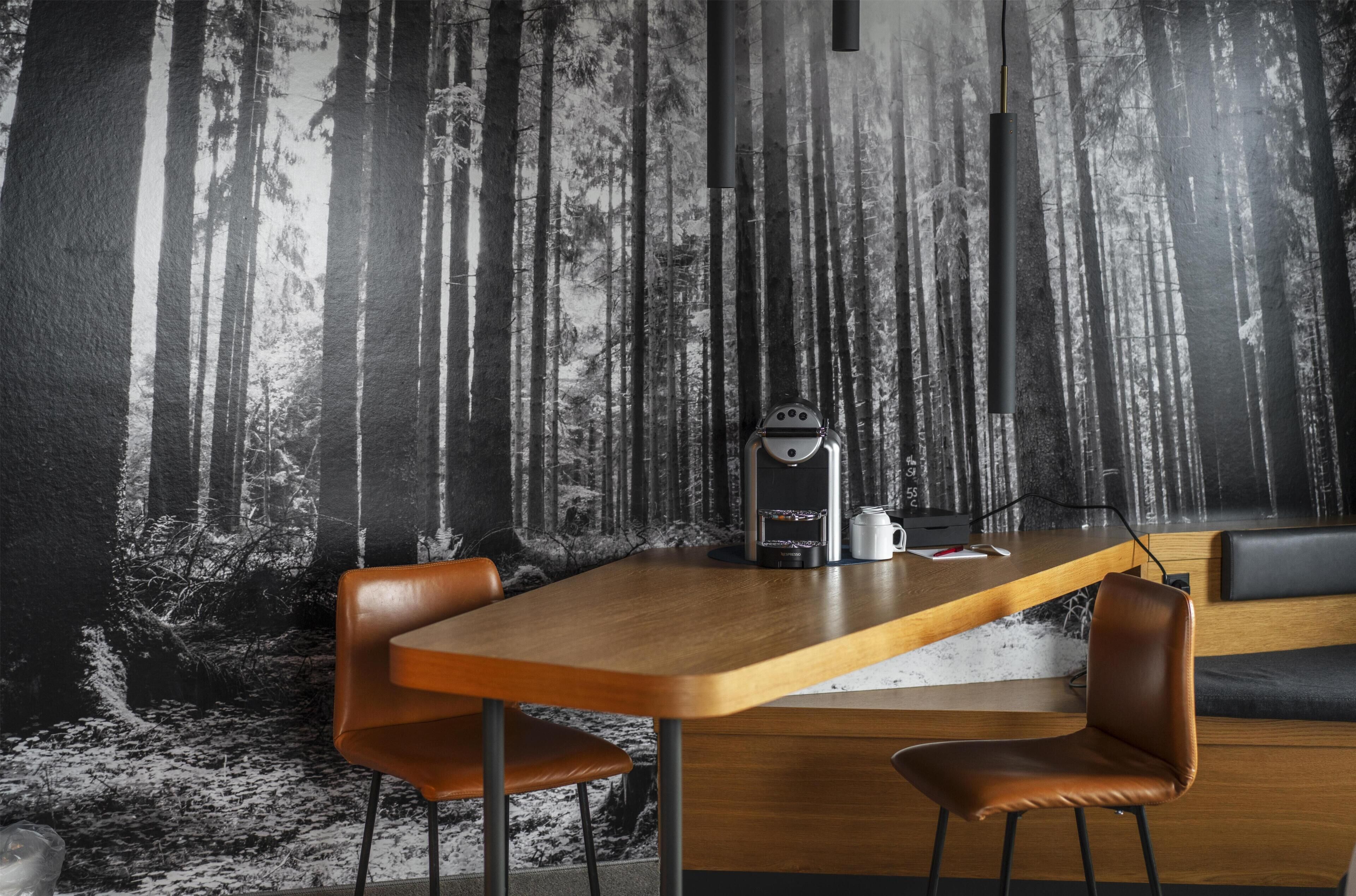 Una habitación moderna con una larga mesa de madera, dos sillones de cuero, una máquina de café y una pared con un mural de un bosque.