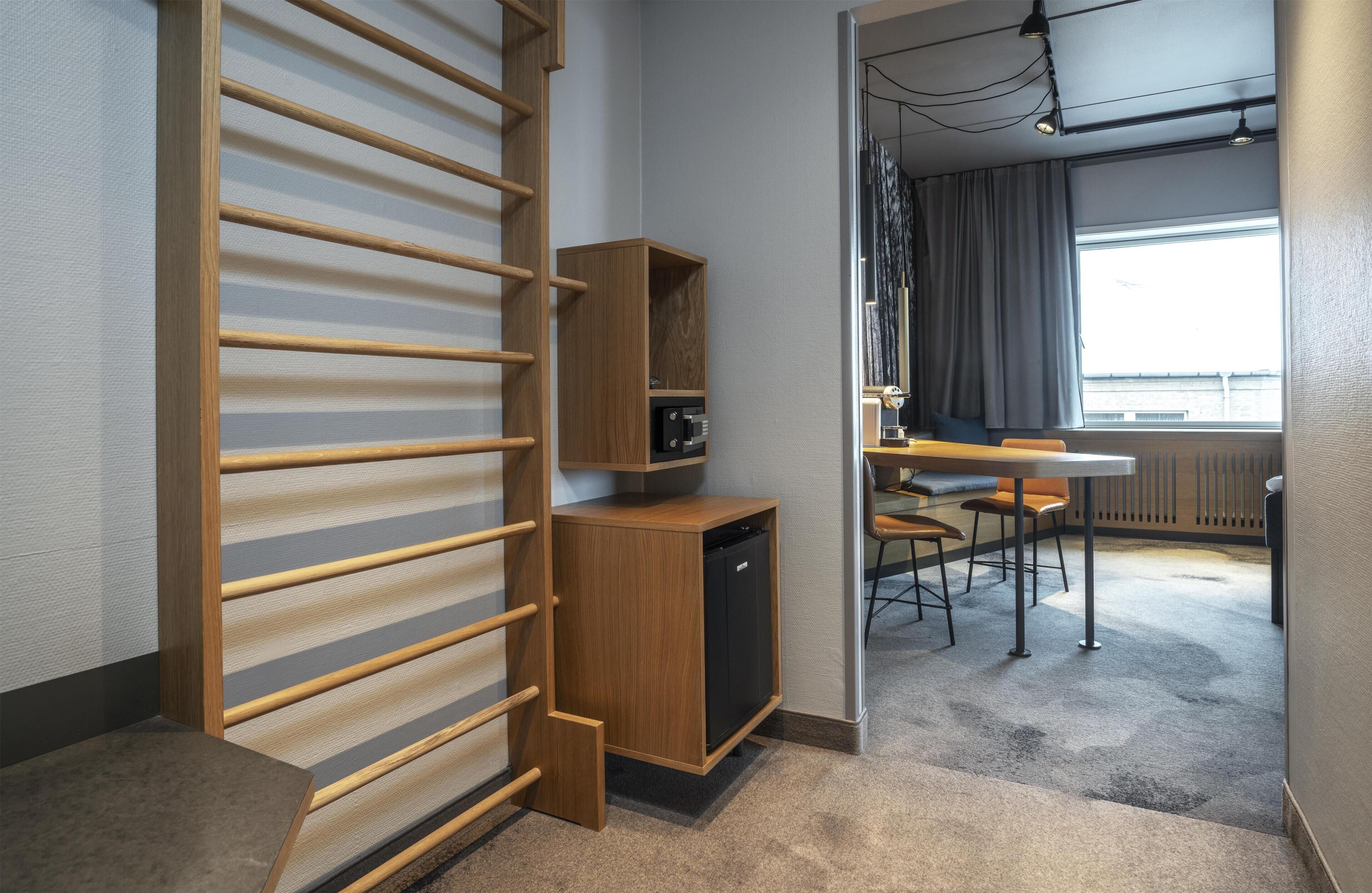 A modern hotel room with a wooden wardrobe, a desk with a chair, and a dining area with a table and chairs.