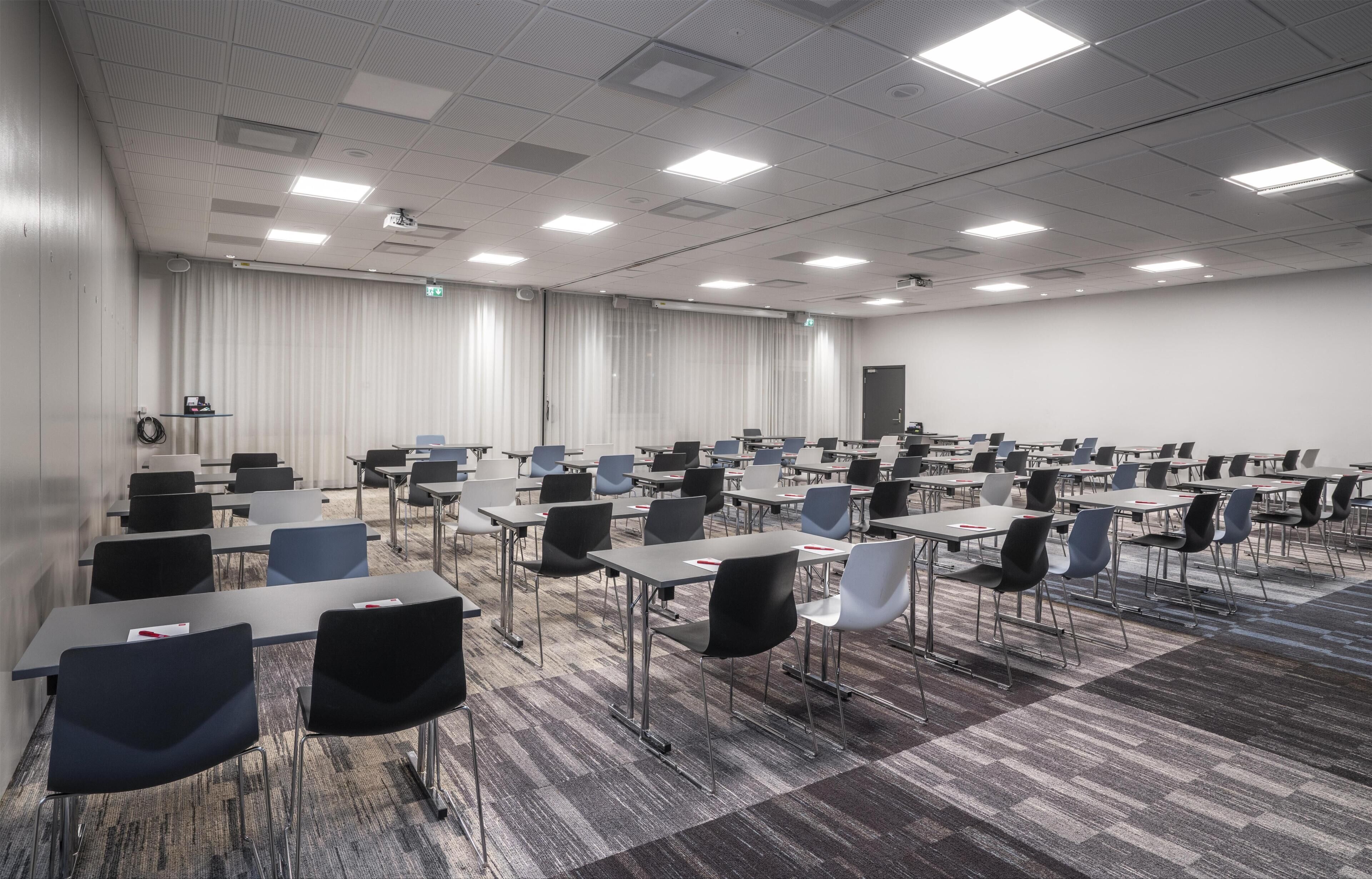Un salón de conferencias con filas de mesas y sillas, un proyector y un ventanal con cortinas.