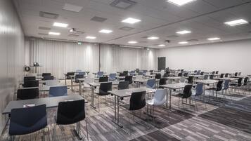 Un salón de conferencias con filas de mesas y sillas, un proyector y un ventanal con cortinas.