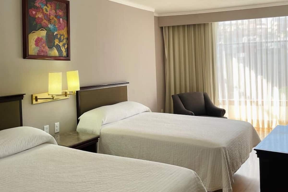 A hotel room with two beds, a desk, a chair, a painting on the wall, and a window with curtains.