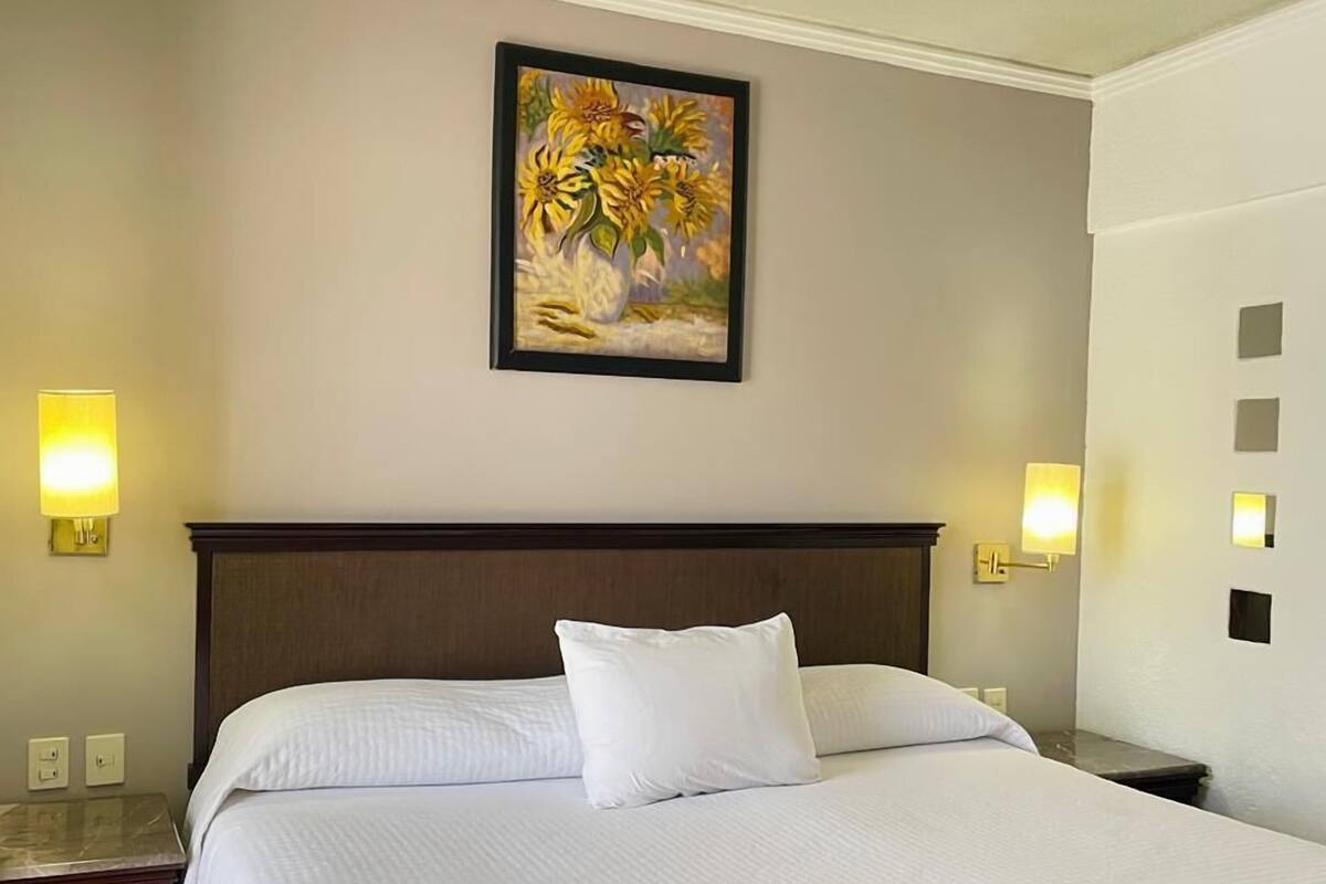 A hotel room with a large bed, two bedside tables, a nightstand, a painting on the wall, and two wall-mounted lamps.