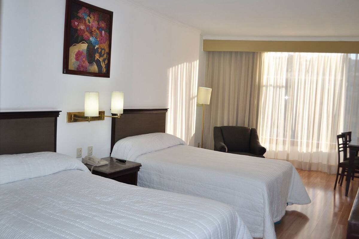 A hotel room with two beds, a painting, a desk with a lamp, and a chair.