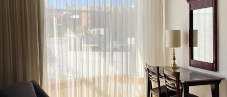 Habitación de hotel con cama, escritorio y silla. Cuenta con un ventanal con cortinas, una lámpara en el escritorio y un espejo en la pared.