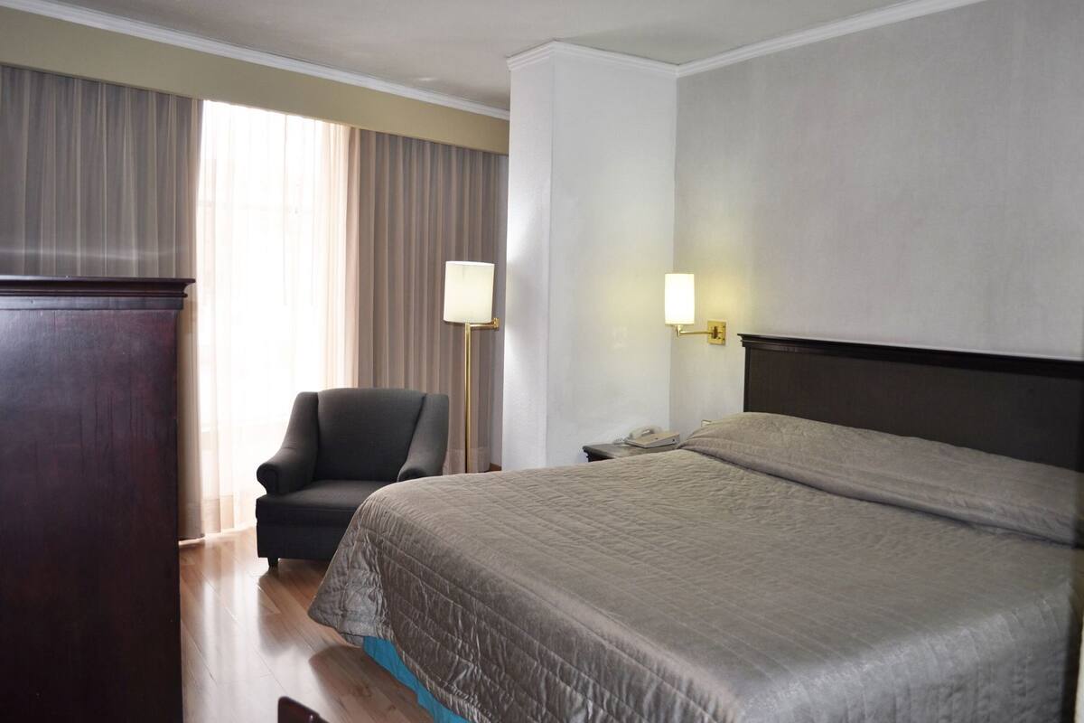 A hotel room with a large bed, a chair, a desk with a lamp, and a telephone.