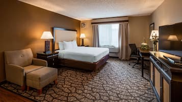 A hotel room with a large bed, two bedside tables with lamps, a desk with a chair, a television, and a patterned carpet.