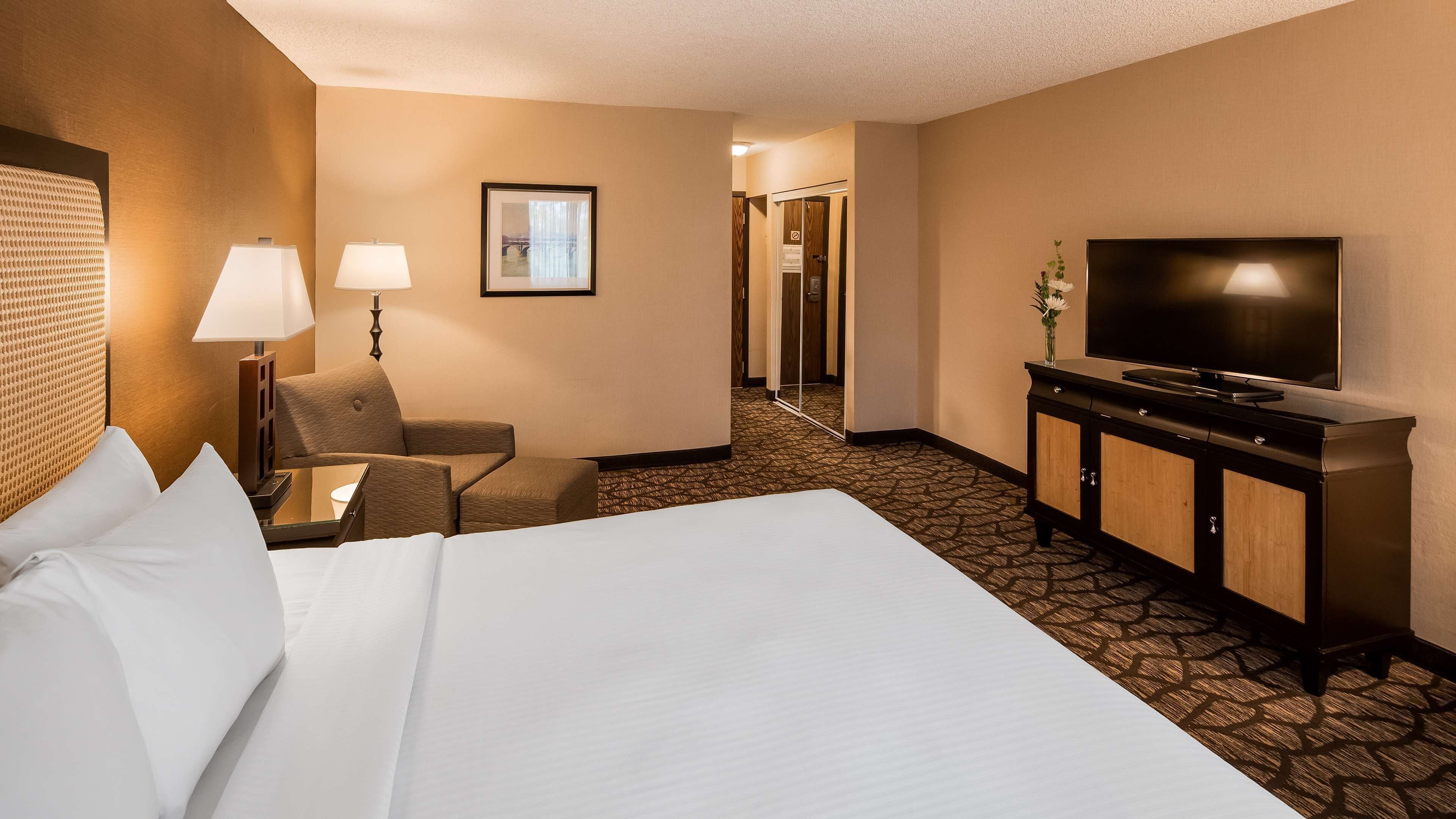 A hotel room with a large bed, a desk with a lamp, a chair, a television, and a framed picture on the wall.