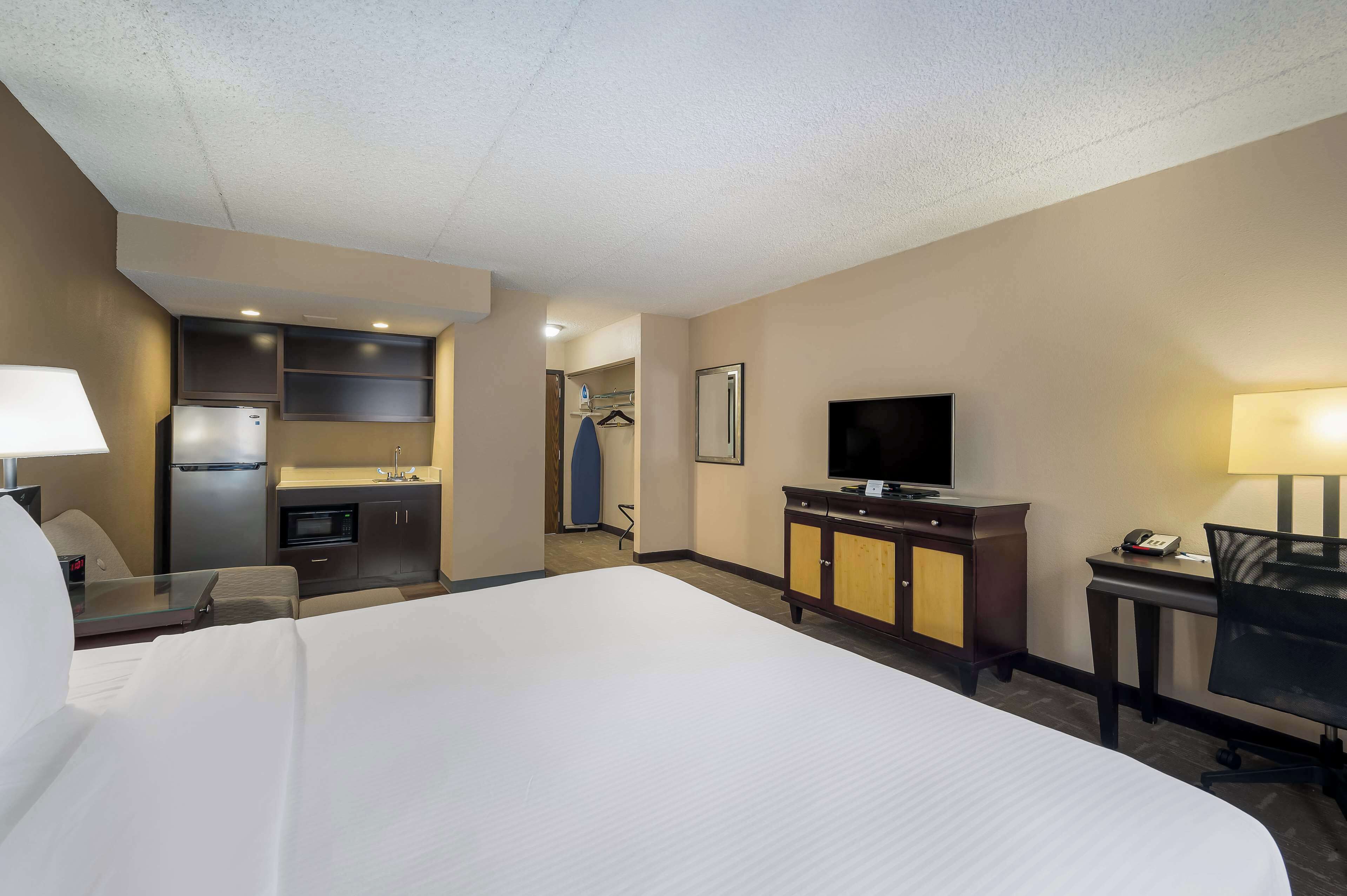 A hotel room with a large bed, a desk with a chair, a television, a minibar, a refrigerator, a microwave, and a small kitchenette.