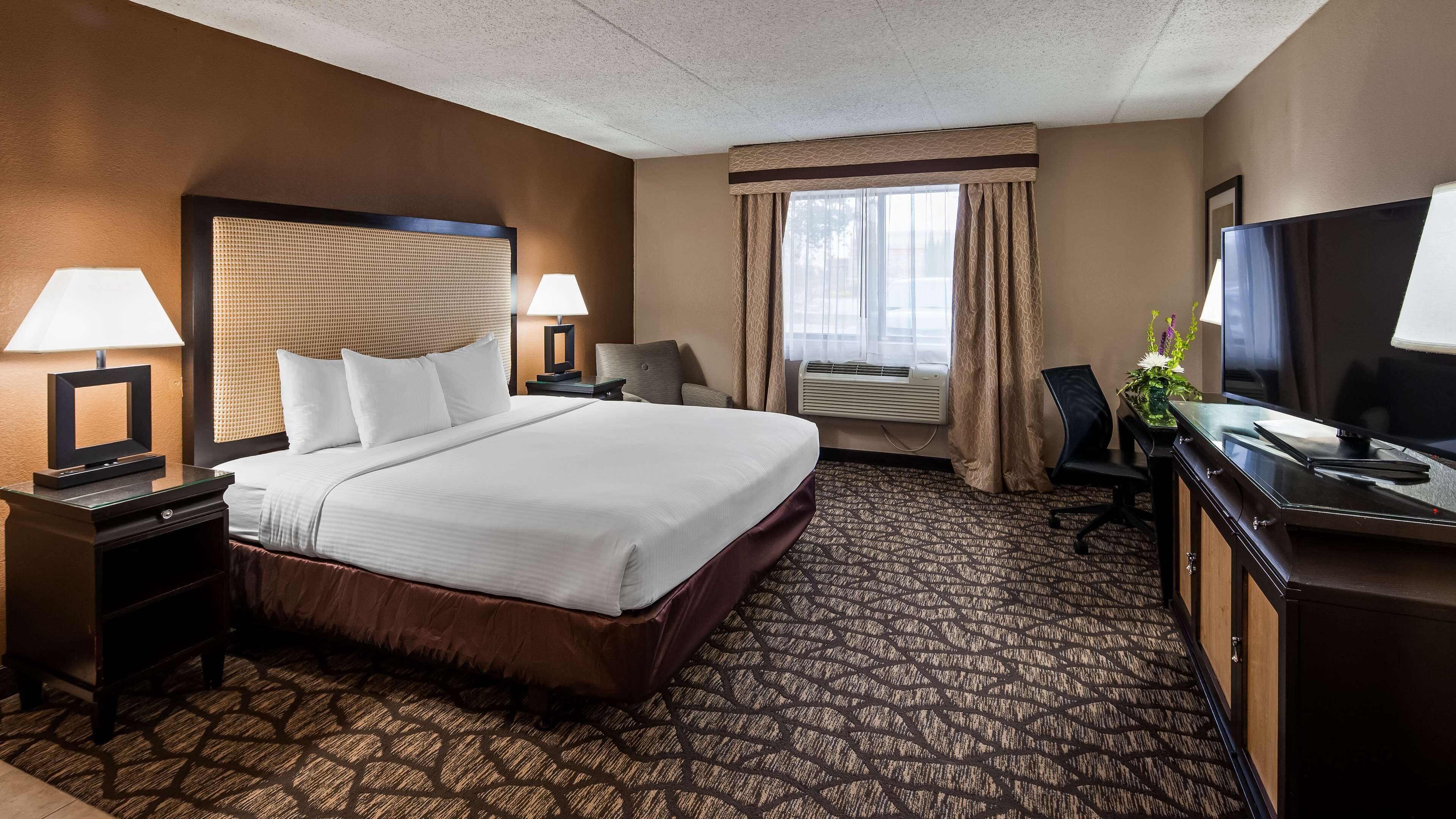 Habitación de hotel con una cama grande, dos mesitas de noche con lámparas, un escritorio con televisor y una ventana con cortinas.