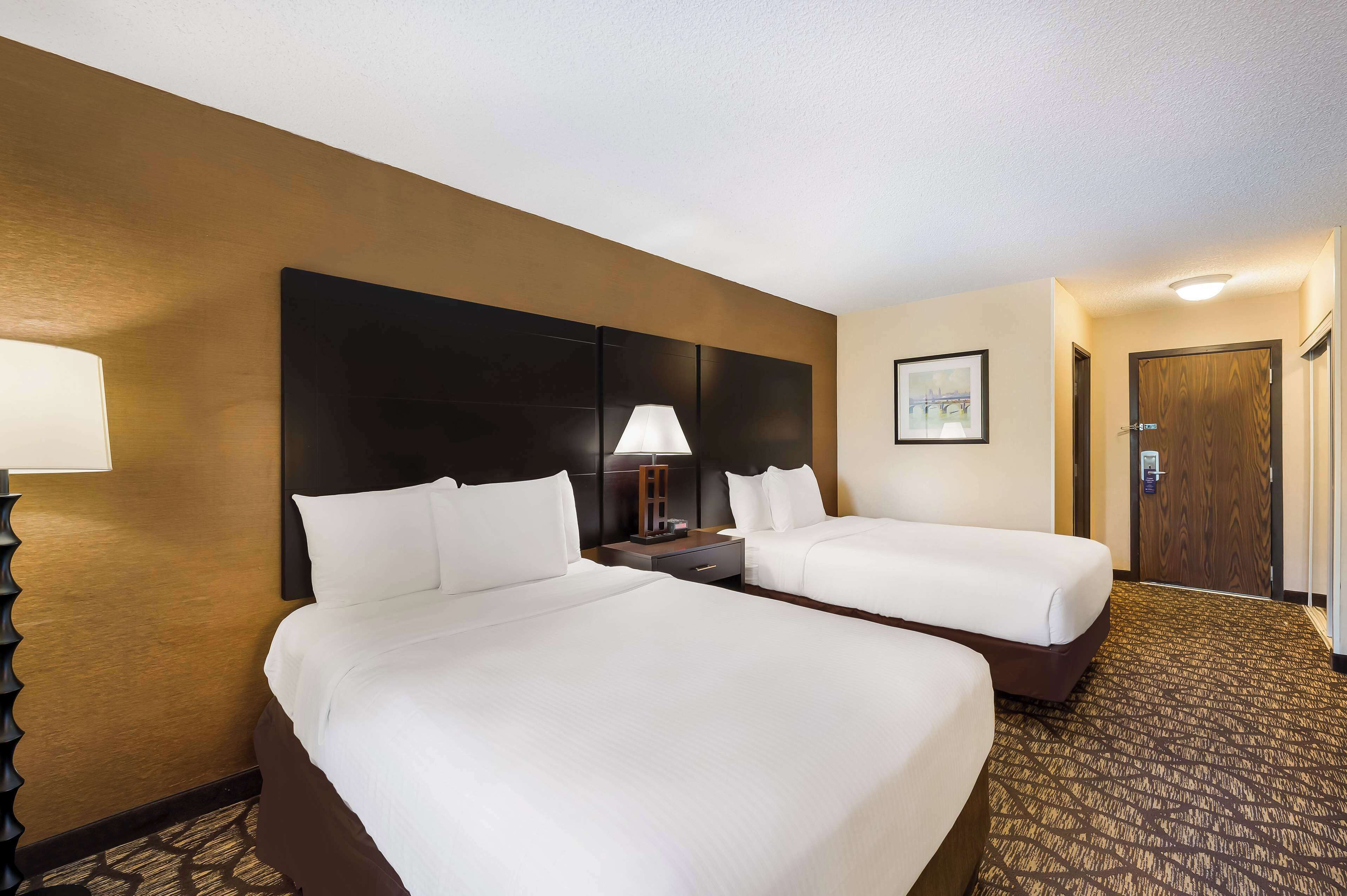 A hotel room with two beds, a nightstand with a lamp, a framed picture on the wall, and a wooden door.