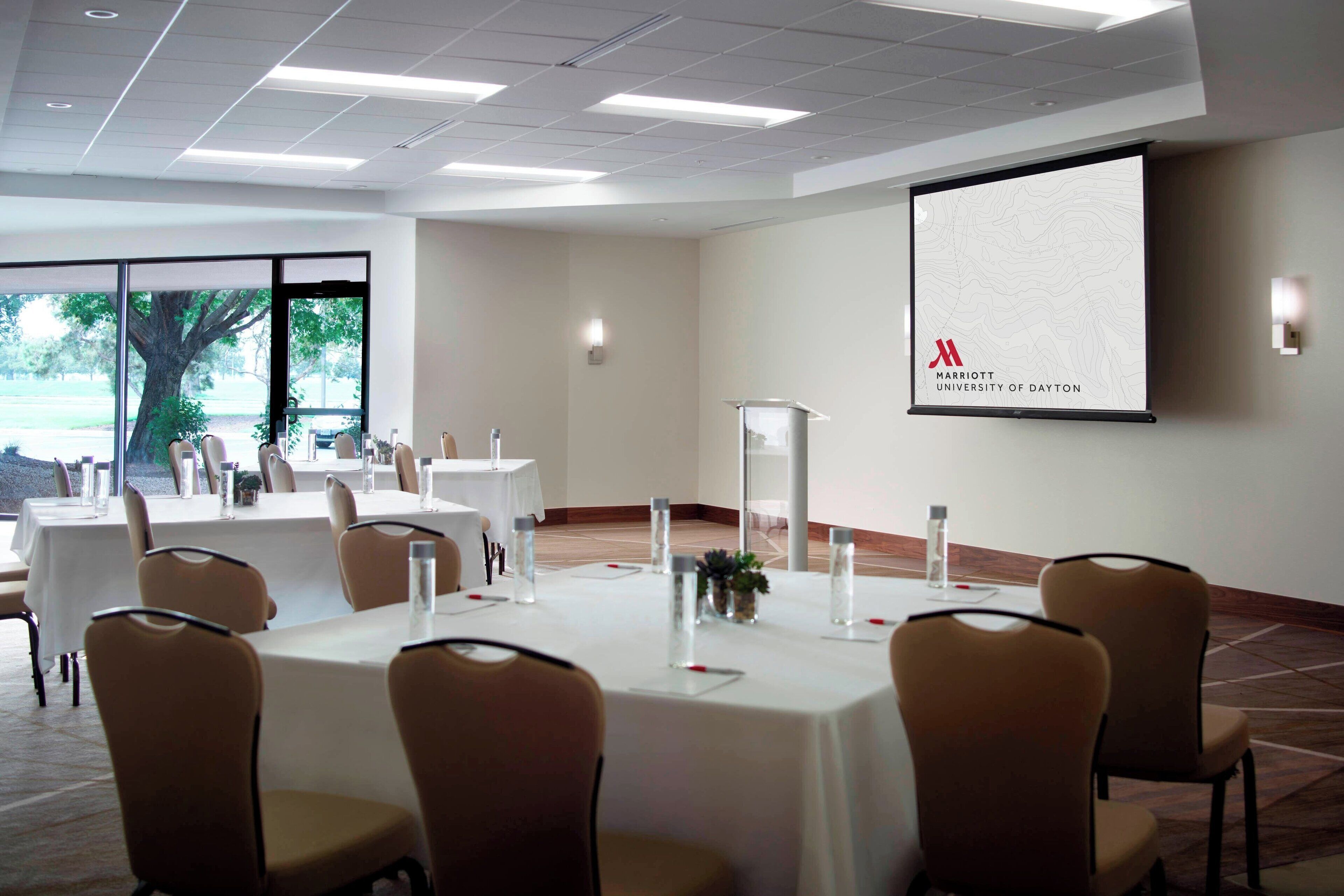 A conference room with tables and chairs, a projector screen displaying the Marriott University of Dayton logo, and a view of trees outside.