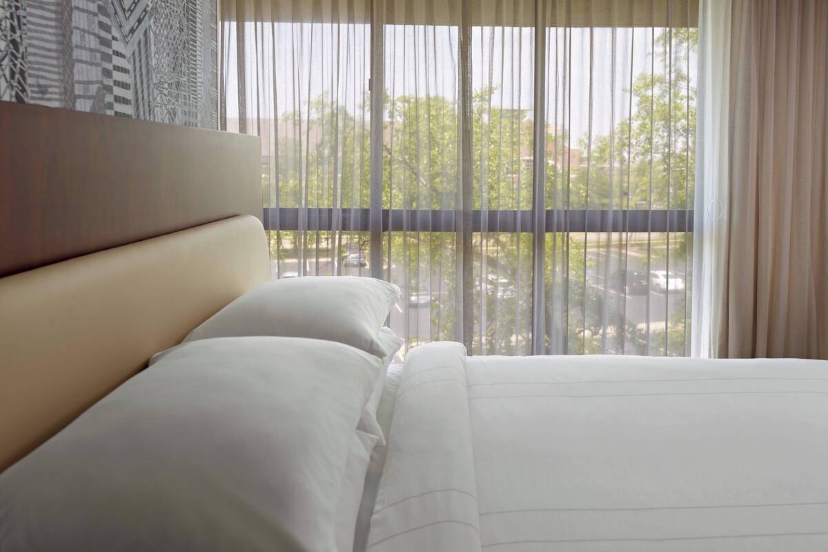 A hotel room with a large bed, a headboard, and a view of a cityscape through sheer curtains.