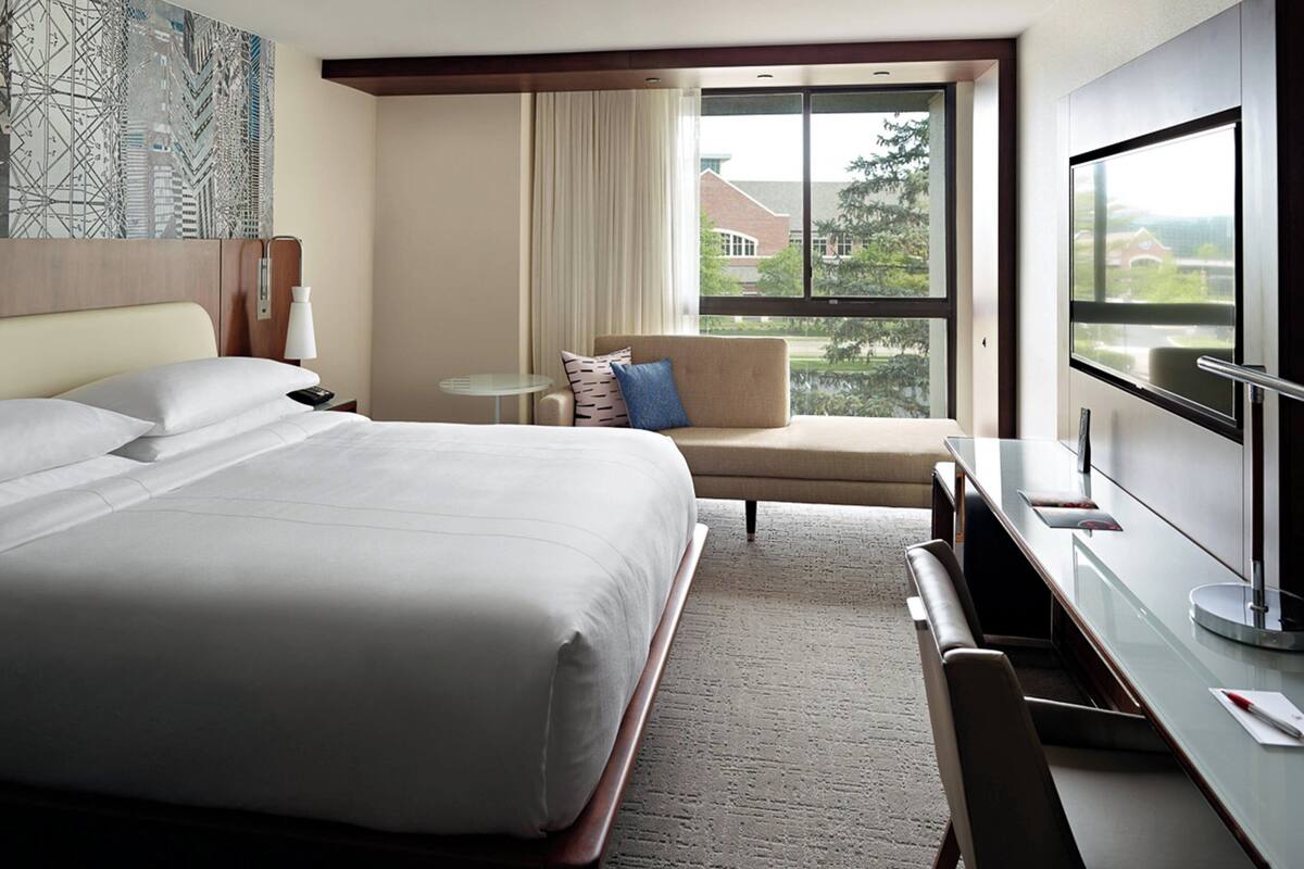 A hotel room with a large bed, a desk, a chair, a TV, and a window with a view of buildings and trees.