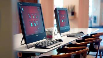 Deux ordinateurs Lenovo sont posés sur une table blanche, accompagnés d’un clavier et d’une souris. On aperçoit également des chaises en bois et une plante en arrière-plan.