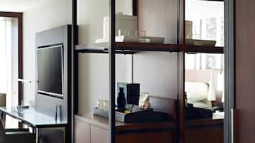 A hotel room with a TV, a desk with a lamp, and shelves with various items.