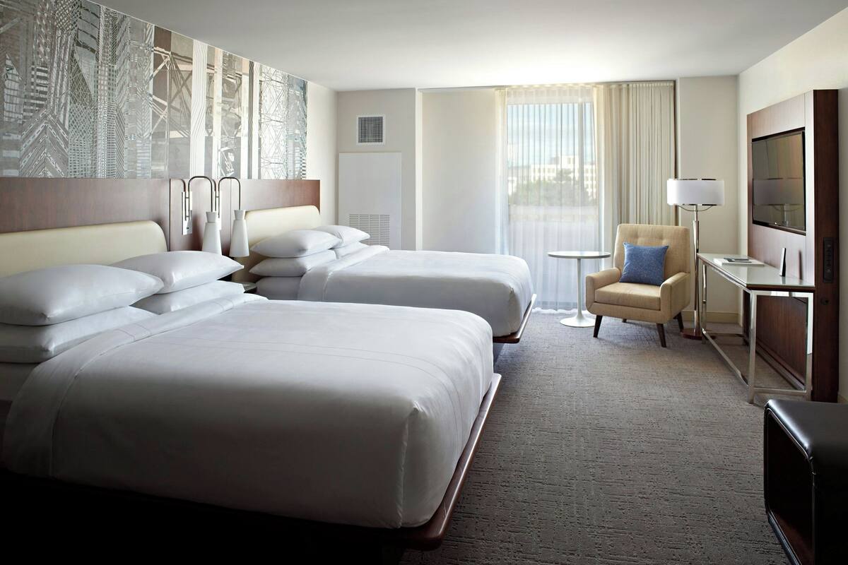A hotel room with two beds, a chair, a TV, and a window with curtains.