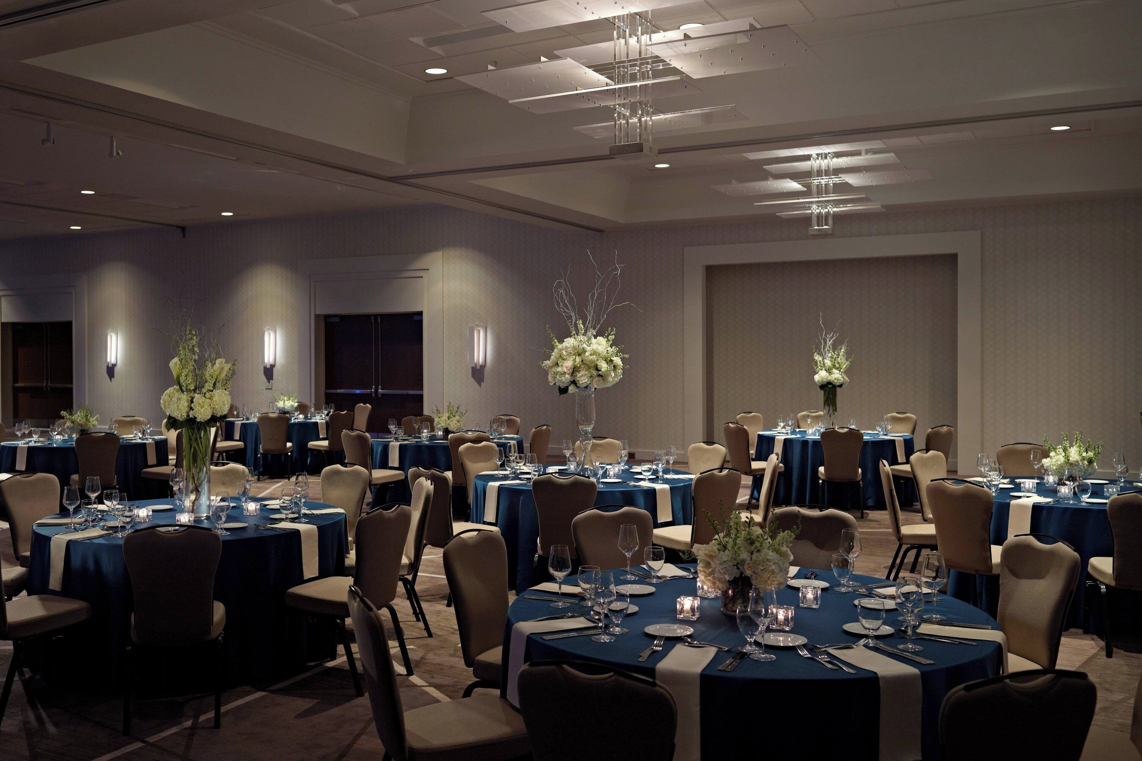 Una sala de banquetes amb taules rodones preparades per dinar, cadascuna amb mantell blau, plats blancs i coberts.