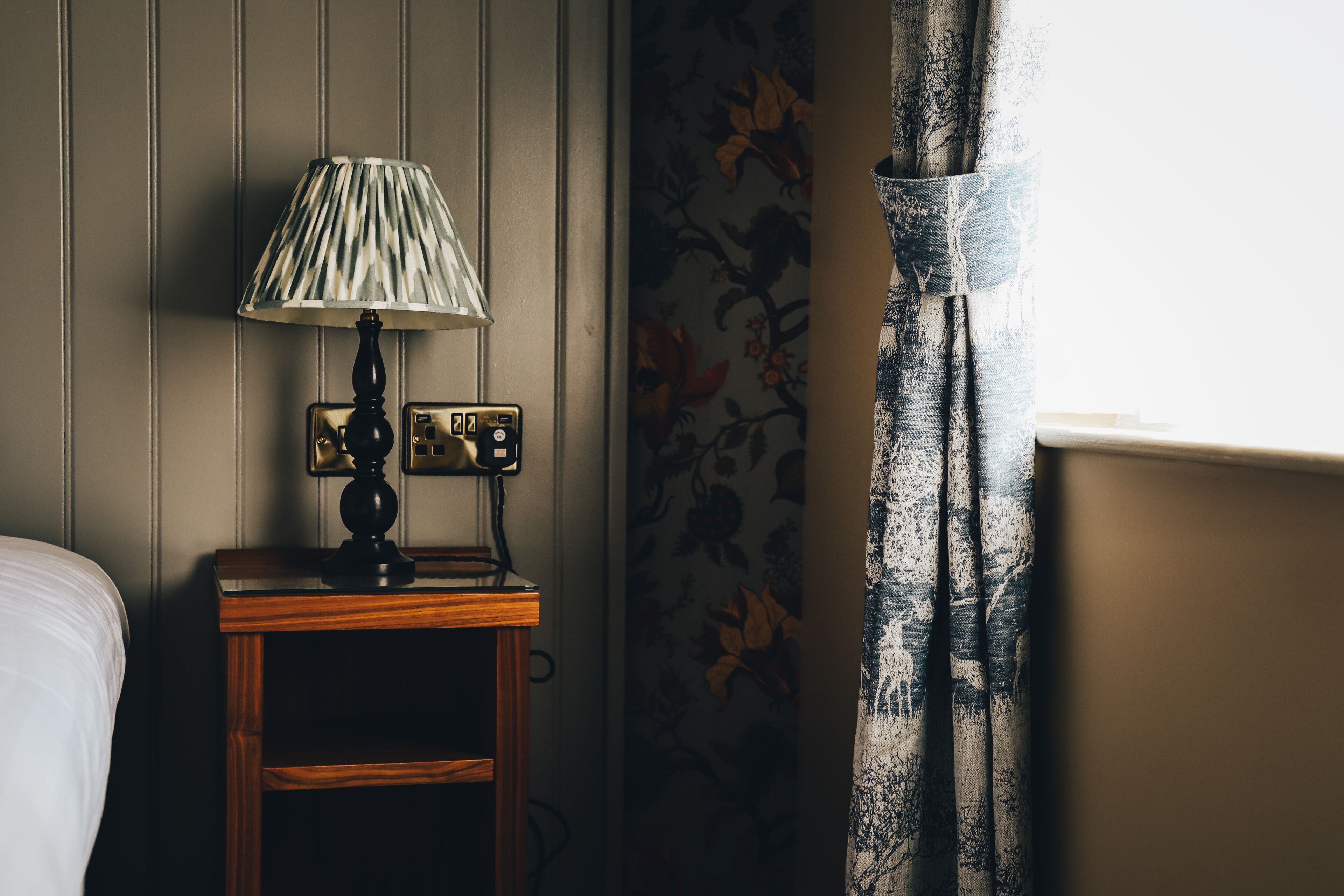 Een houten nachtkastje met een lamp, een stopcontact en een wandcontactdoos.