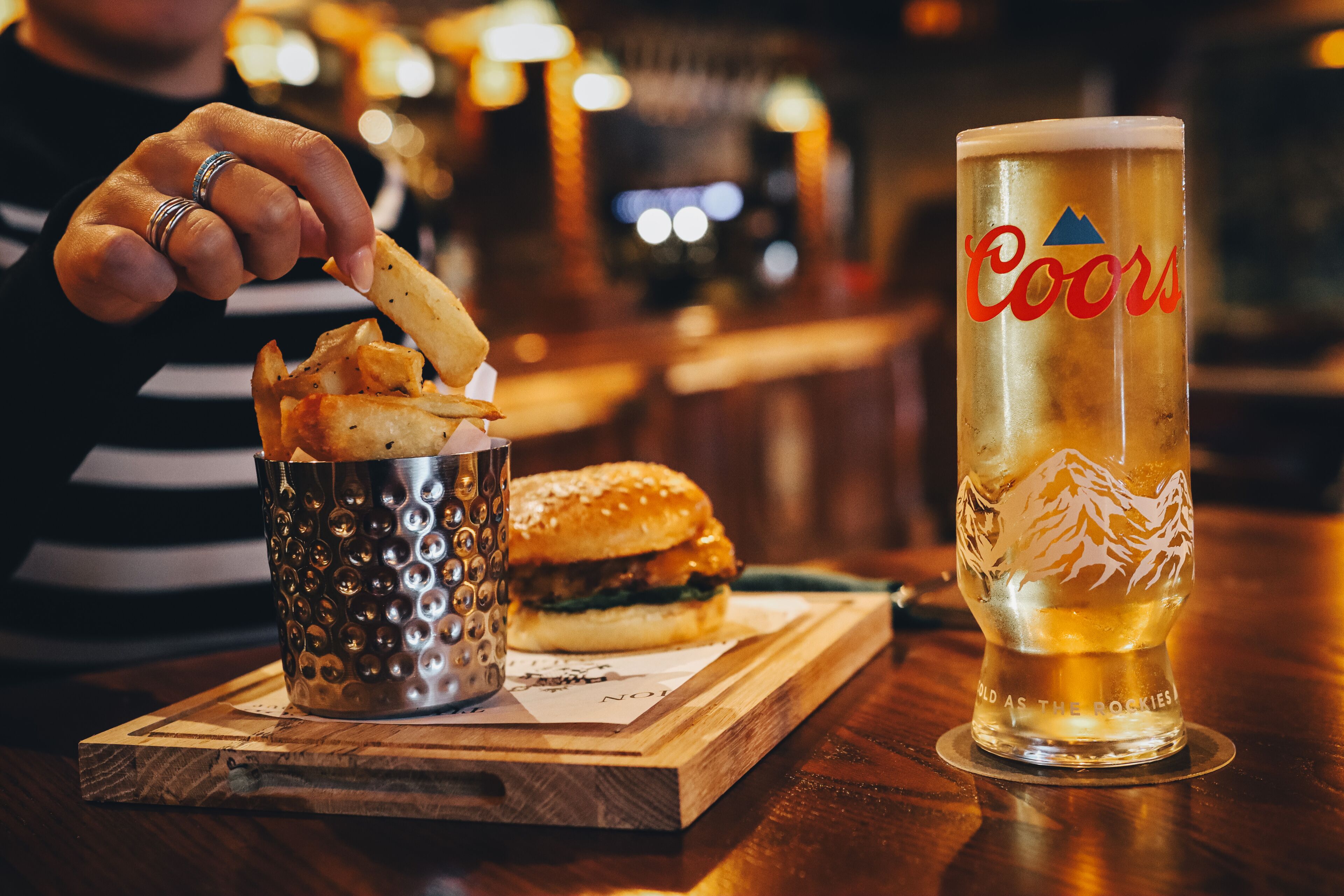 A person dipping fries into a metal cup, a burger on a wooden board, and a tall glass of beer with 'Coors' branding.