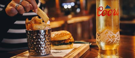 Una persona mojando papas fritas en un vaso metálico, una hamburguesa sobre una tabla de madera y un vaso alto de cerveza con la marca 'Coors'.