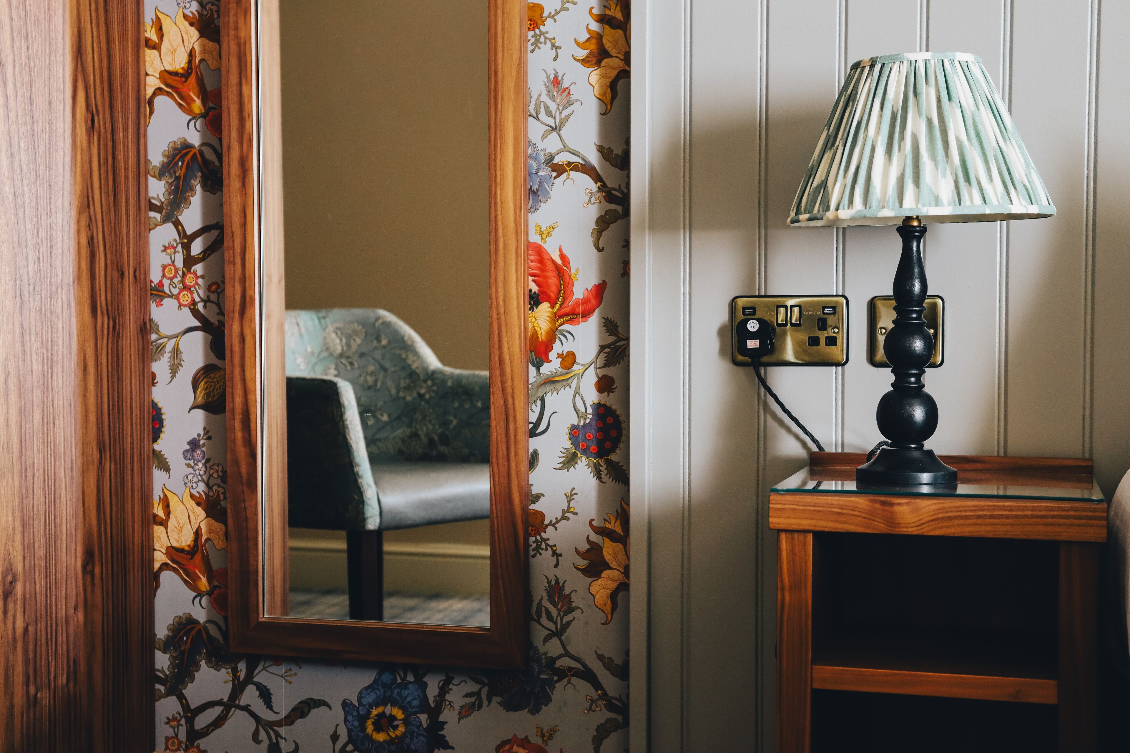 Un miroir à cadre en bois, un papier peint à motifs floraux, une lampe posée sur une table de chevet en bois et une prise électrique.