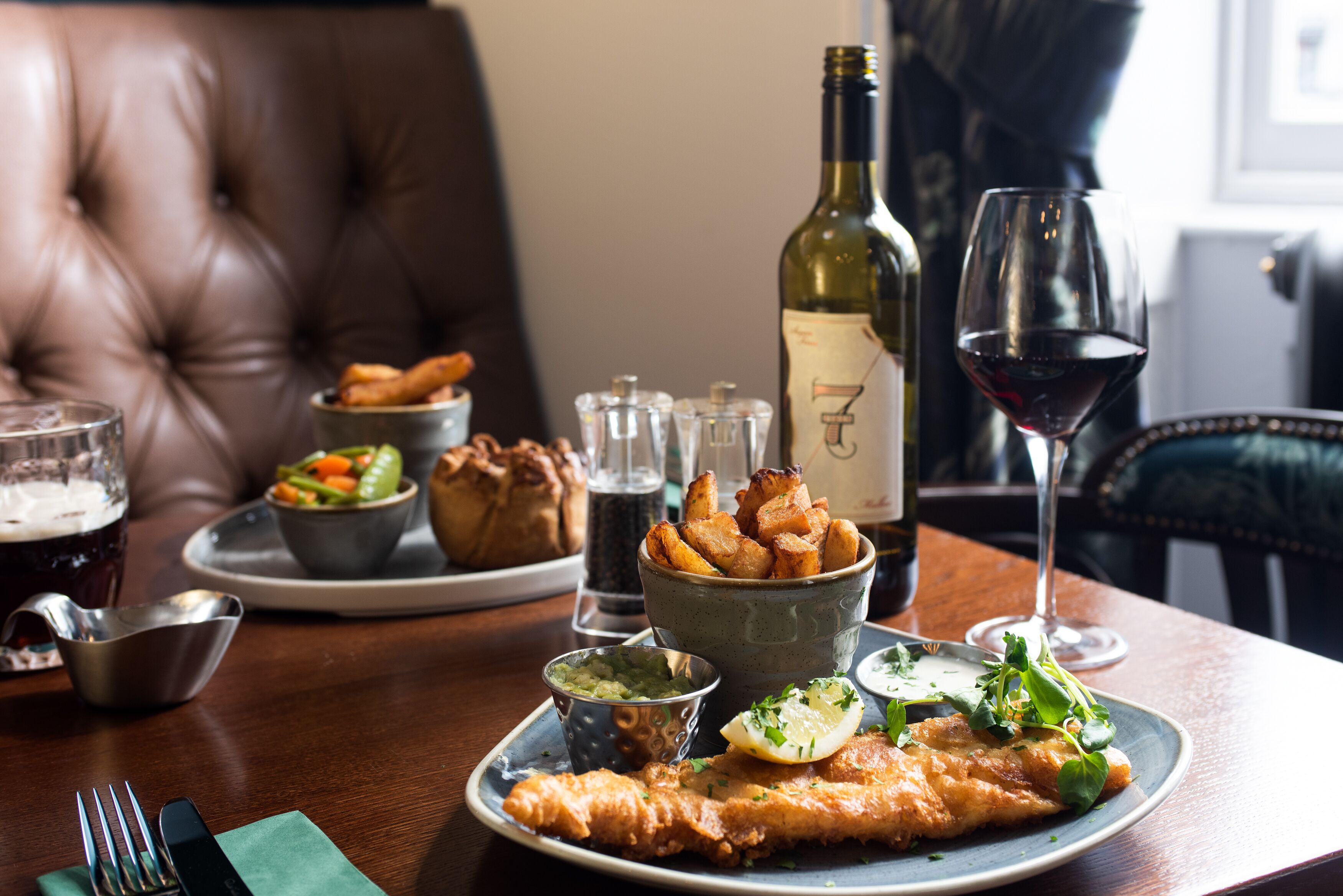 A dining table with a plate of fish and chips, a bottle of wine, and various side dishes.