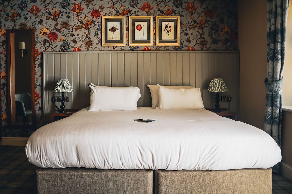 Un grand lit avec deux oreillers, une table de chevet avec une lampe et un tableau floral encadré accroché au mur.