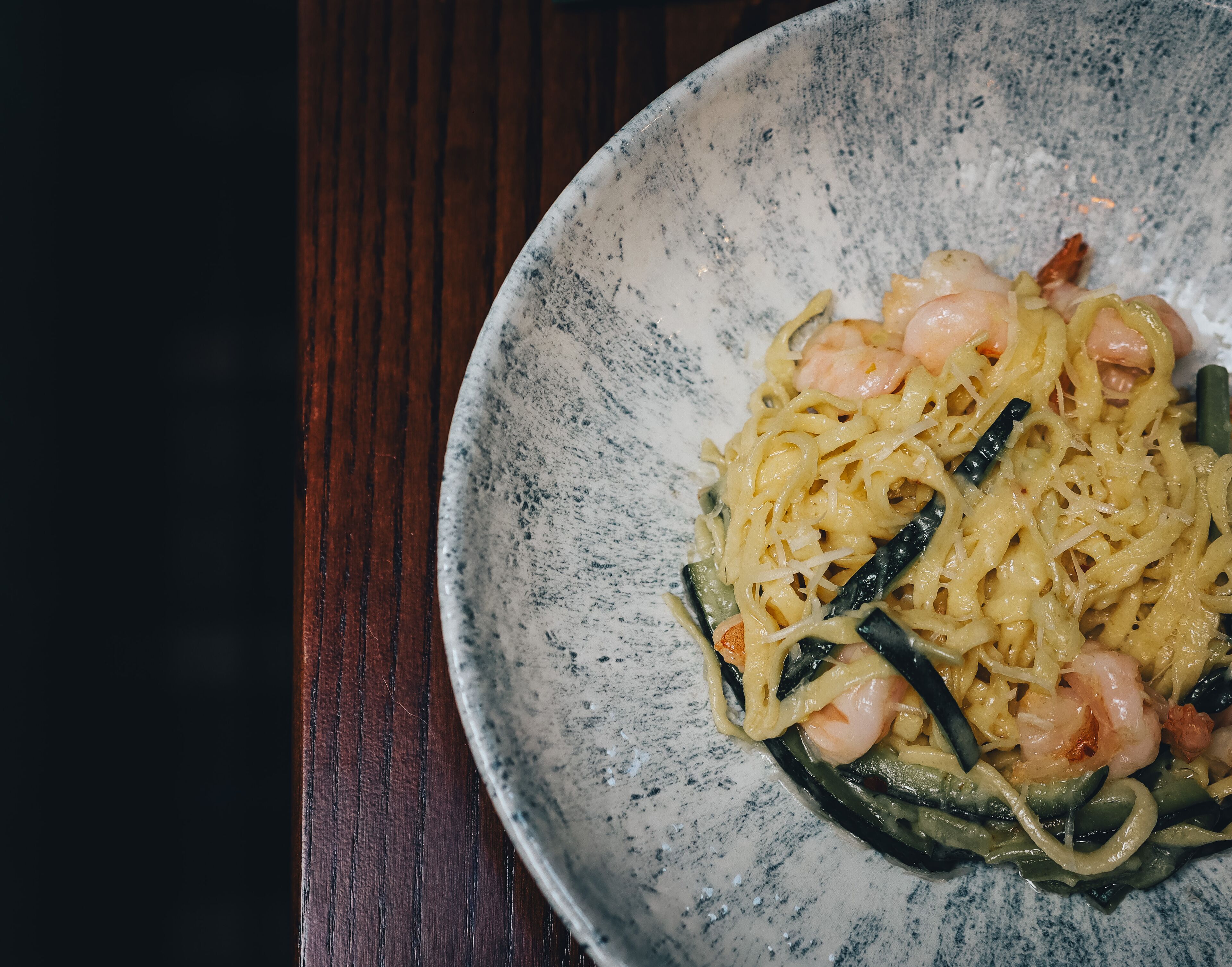 A bowl of pasta with shrimp and vegetables on a speckled ceramic plate.