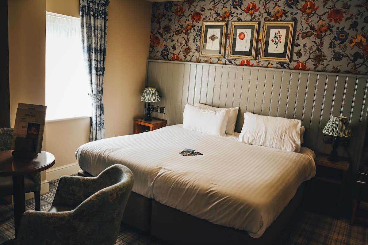 Une chambre d’hôtel avec un grand lit, deux tables de chevet avec des lampes, une chaise et un papier peint à motifs floraux.