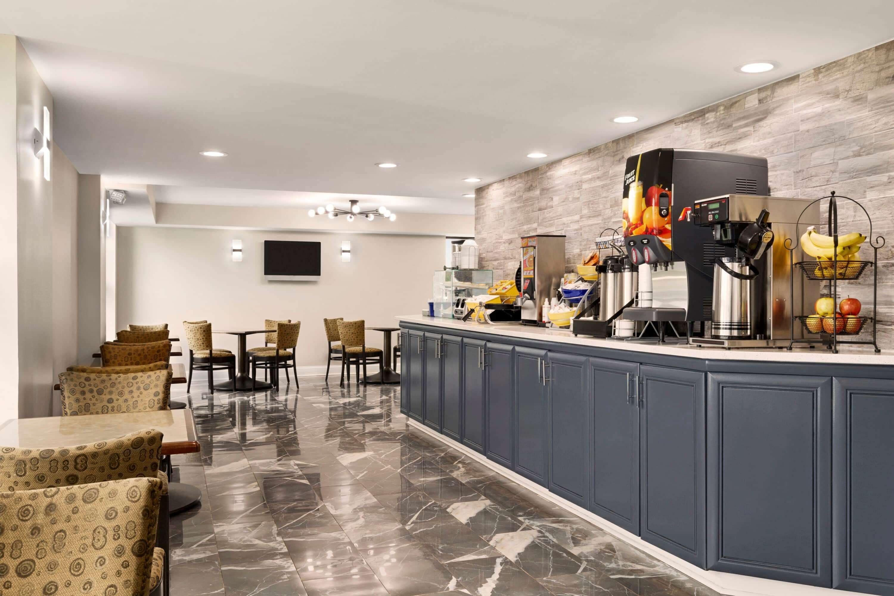 Un hall d’hôtel moderne avec un sol en marbre, un espace salon doté de fauteuils à motifs, un comptoir avec une machine à café et un panier de fruits, et un téléviseur à écran plat fixé au mur.