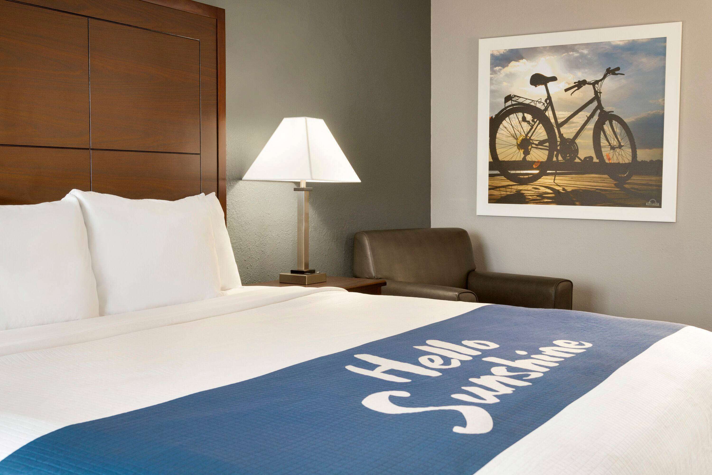 A hotel room with a bed, a chair, a lamp, and a wall art of a bicycle.