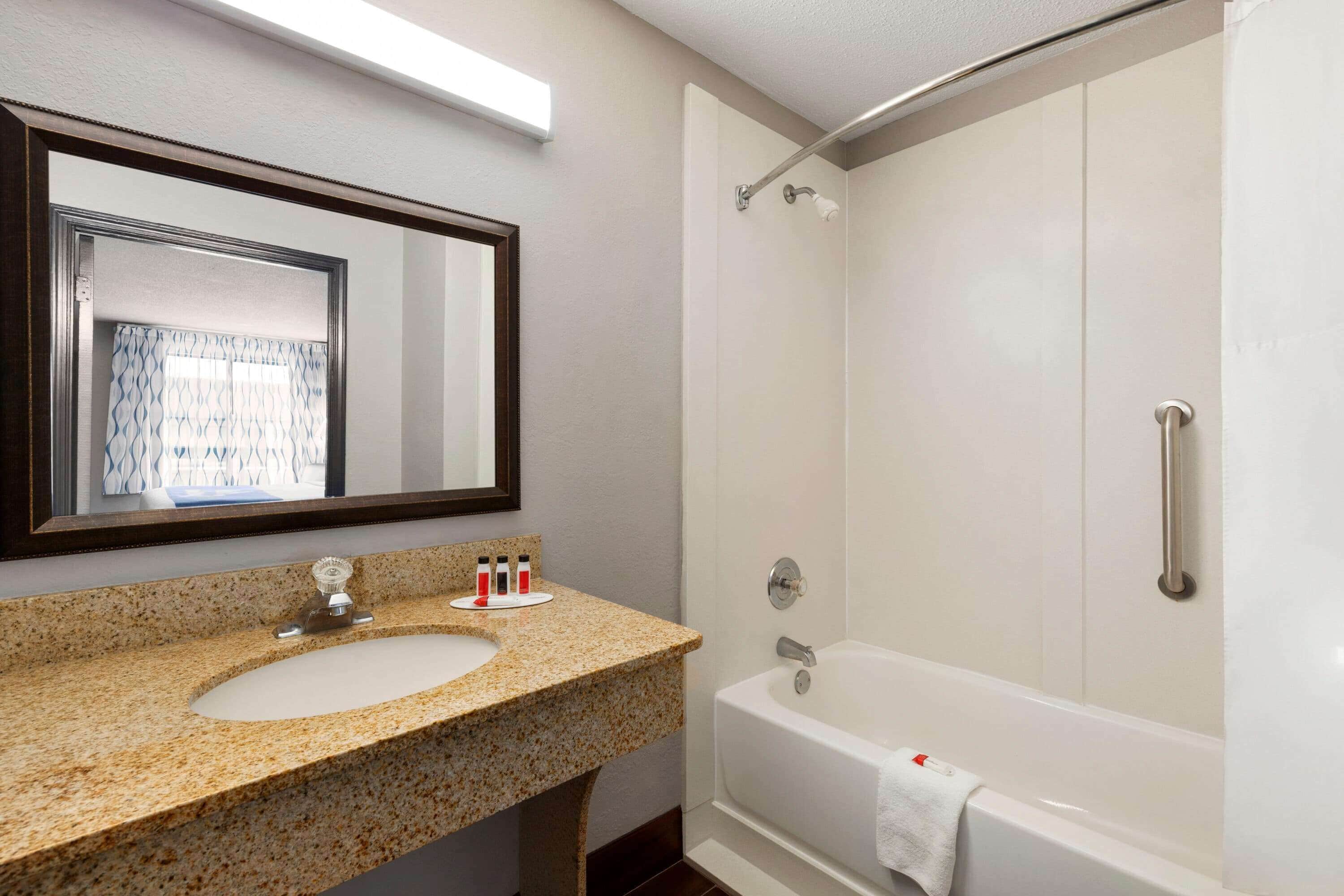 A bathroom with a granite countertop sink, a mirror, a shower with a handheld showerhead, and a white bathtub.