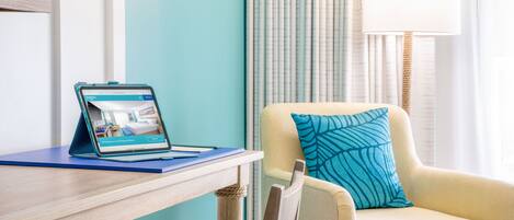 A hotel room with a wooden desk, a laptop displaying a room view, a chair with a blue pillow, and a window with curtains.