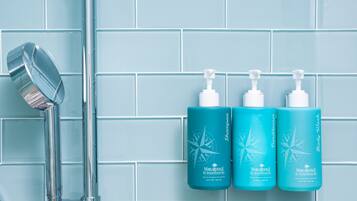 A bathroom with a blue tiled wall, a chrome showerhead, and three bottles of shampoo and conditioner.