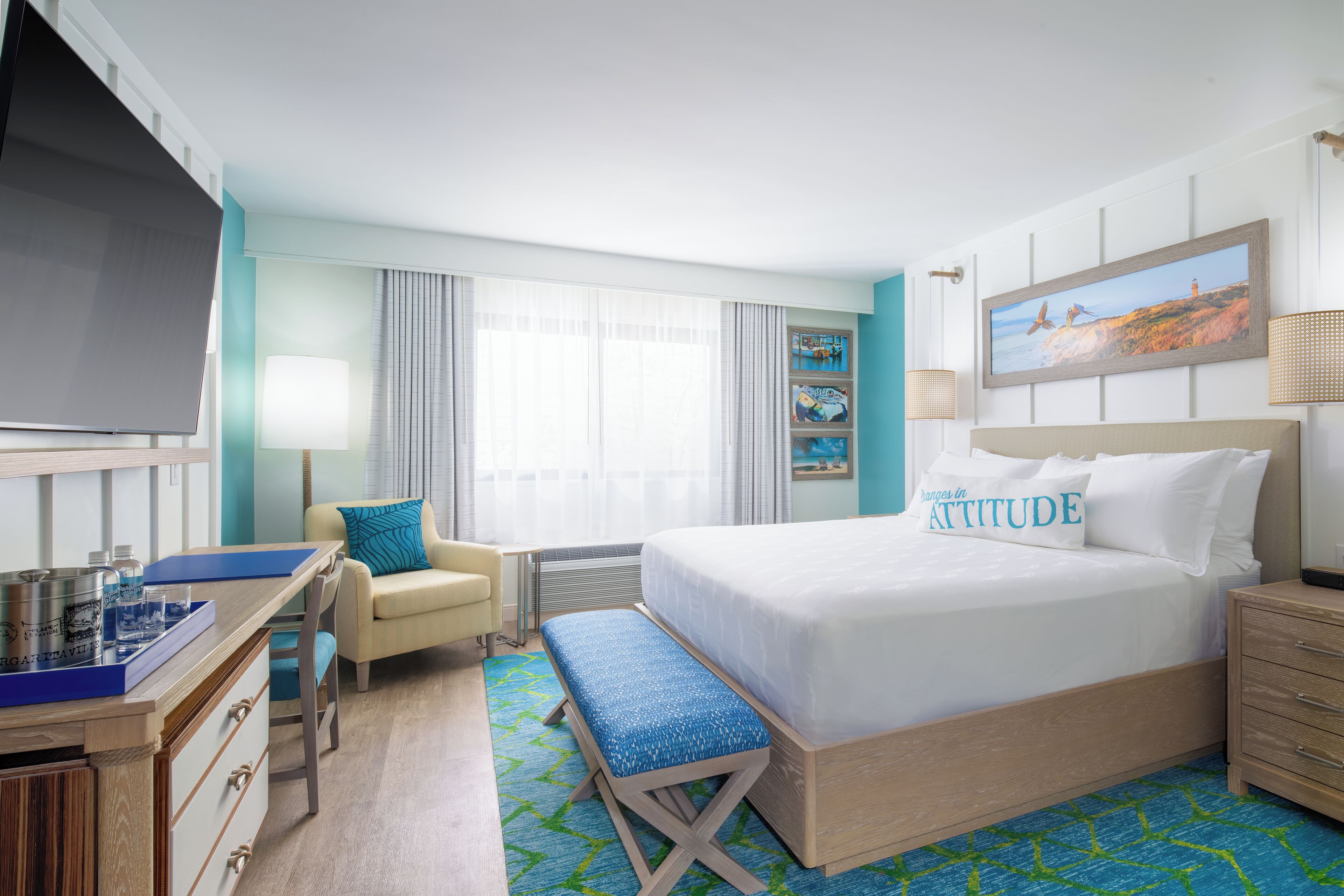 A hotel room with a bed, a desk with a computer, a chair, a bench, a TV, and a window with curtains.