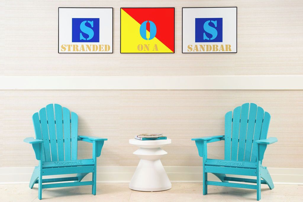 Deux chaises Adirondack turquoise sont placées face à face, avec une petite table blanche posée entre elles. Au-dessus des chaises, trois panneaux encadrés portant les lettres « S », « O » et « S » sont accrochés à un mur en bois clair.