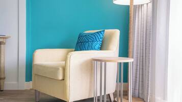 Quarto com parede verde-água, uma poltrona bege com almofada estampada em azul, uma mesa redonda pequena e um abajur.