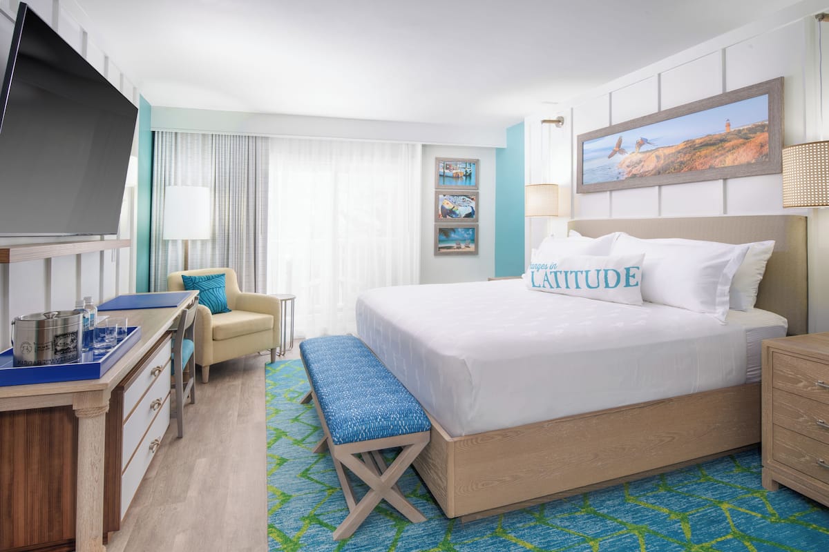 A hotel room with a large bed, a desk with a computer, a chair, and a bench.