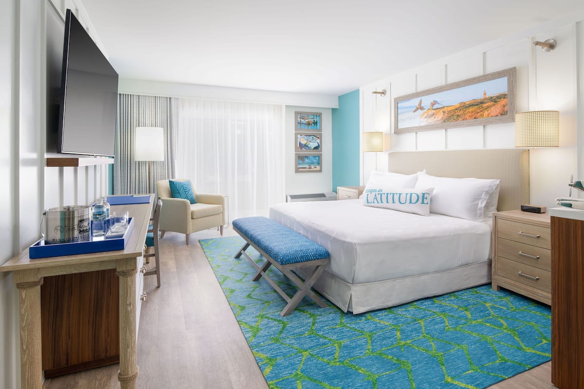 A hotel room with a bed, a desk with a flat-screen TV, a chair, a bench, and a window with curtains.