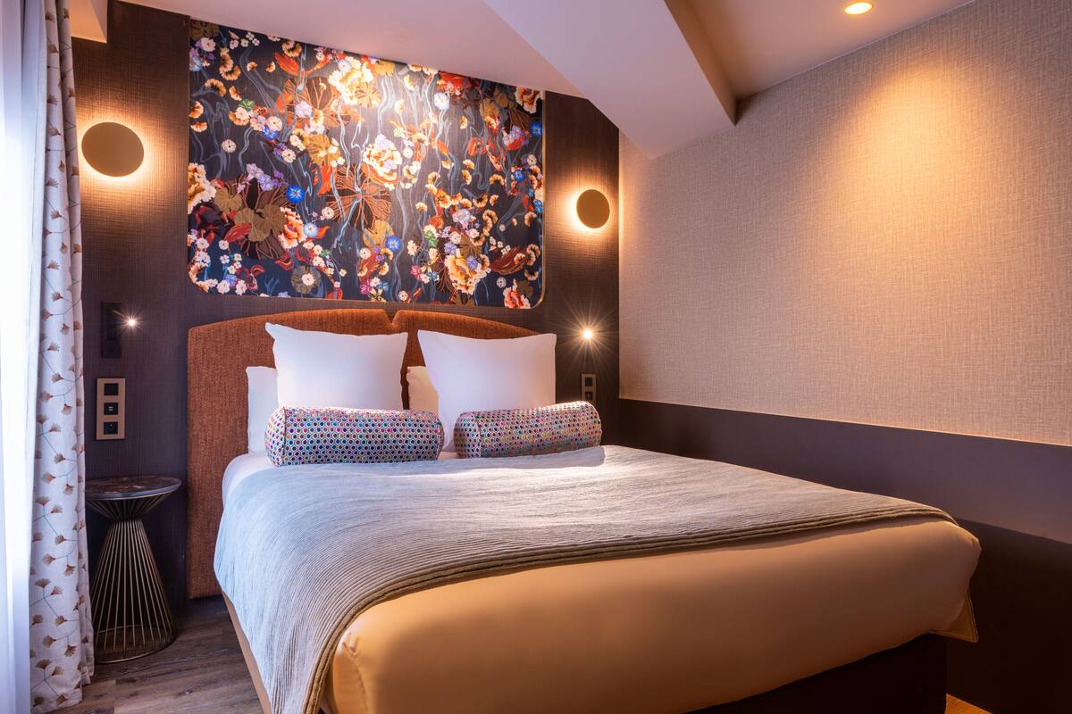 A modern hotel room with a large bed, a decorative wall mural, and two wall-mounted lights.