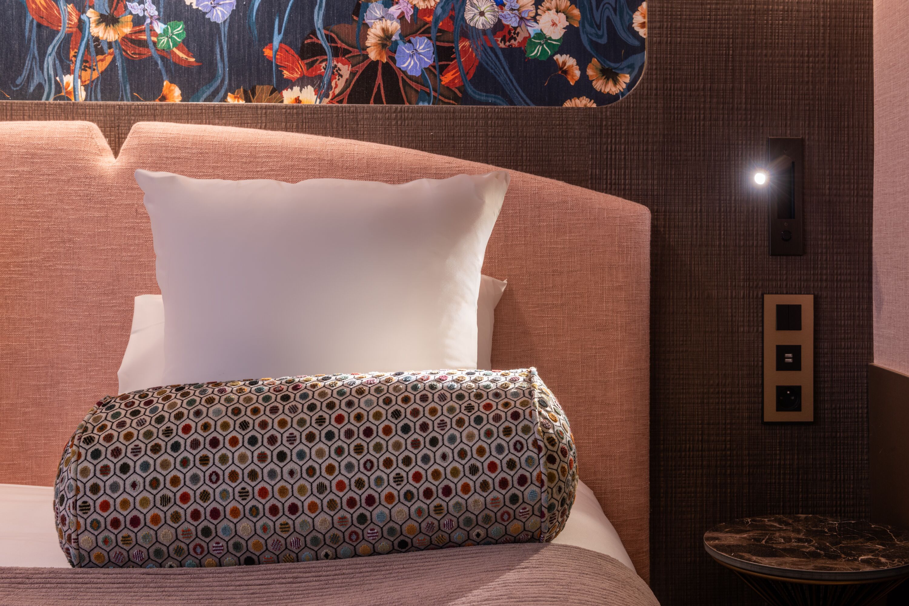 A neatly made bed with a patterned pillow and a plain pillow, a wall-mounted light switch, and a small round table with a dark surface.