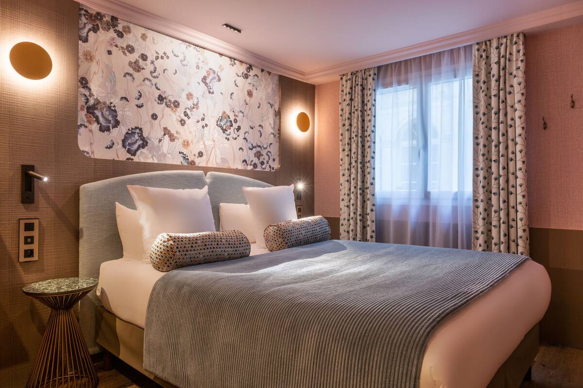 A hotel room with a large bed, floral wallpaper, a round wall light, and a window with sheer curtains.