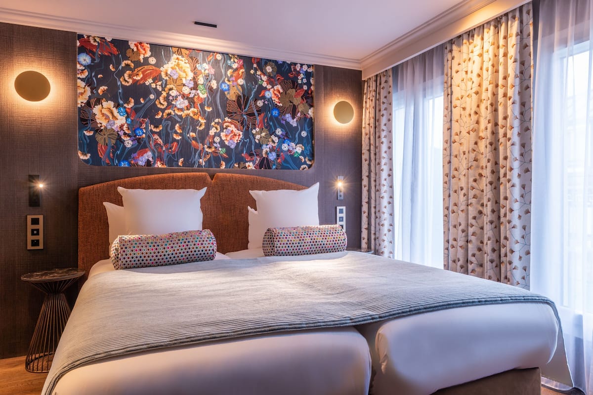 A large bed with a patterned headboard, a wall-mounted artwork, and a view of the outdoors through sheer curtains.