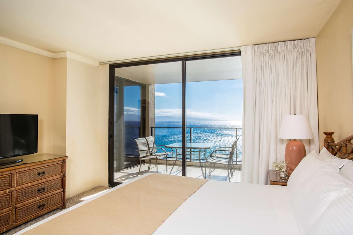 A hotel room with a large bed, a television on a wooden dresser, a balcony with a table and chairs, and a view of the ocean.