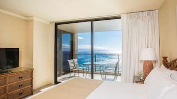 A hotel room with a large bed, a television on a wooden dresser, a balcony with a table and chairs, and a view of the ocean.