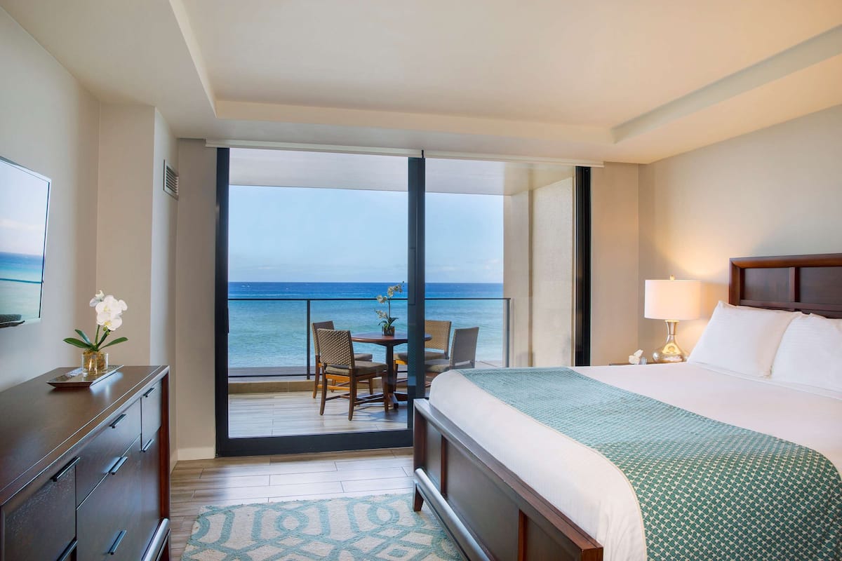 A hotel room with a bed, a desk, a chair, a small table, and a view of the ocean.