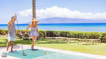 A couple playing shuffleboard on a green court with a view of the ocean and mountains in the background.