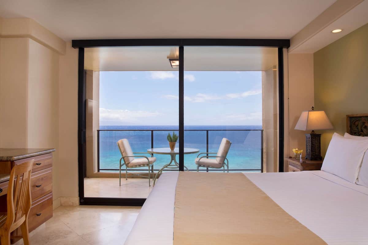 A hotel room with a large bed, a desk, a chair, a small table with chairs, and a balcony with a view of the ocean.
