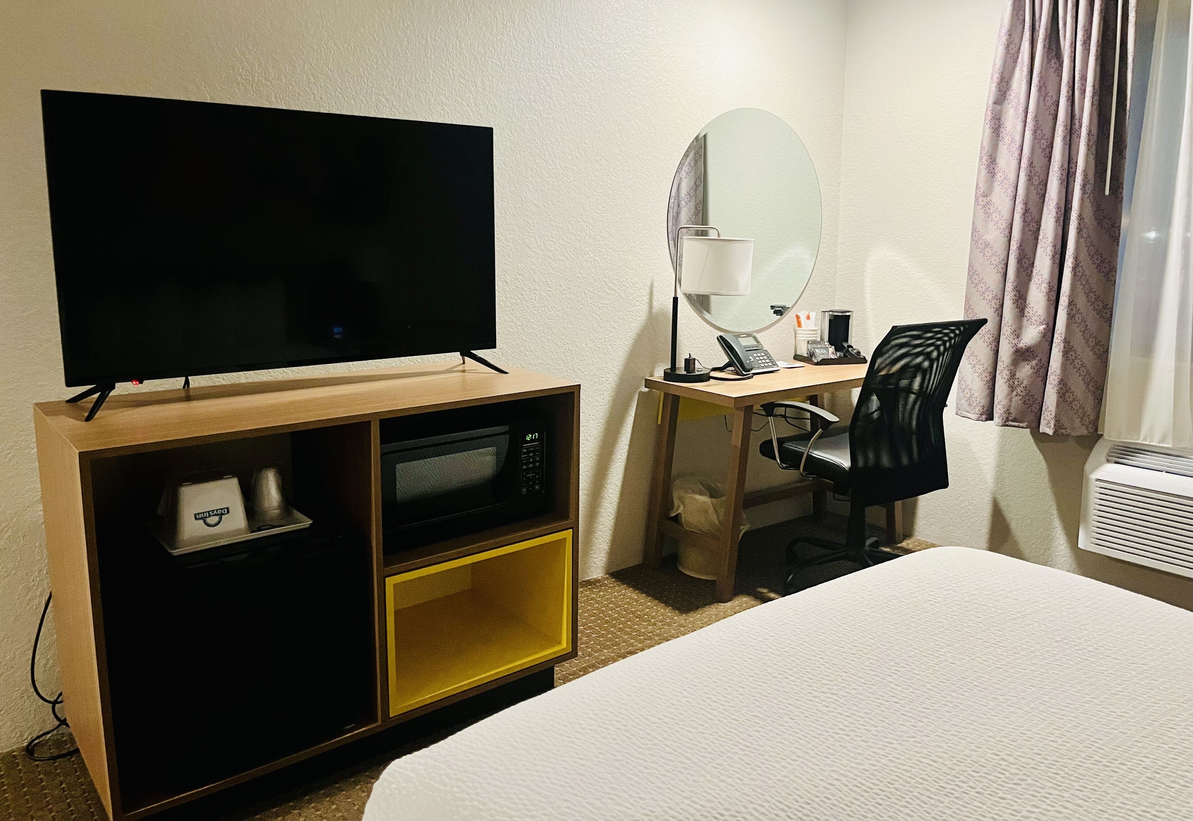 A hotel room with a bed, a TV on a wooden stand, a desk with a lamp, a chair, and a microwave.
